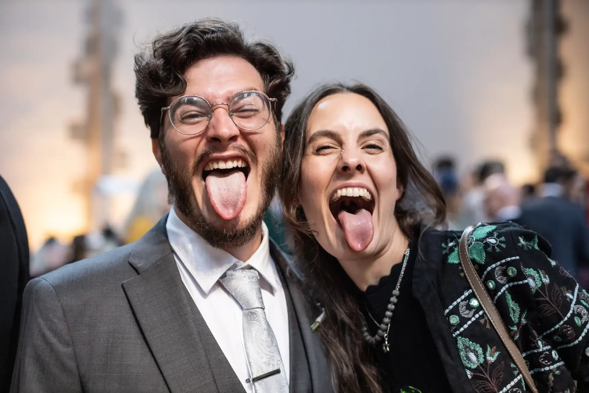 A man and a woman in formal attire playfully stick out their tongues at a social event, smiling at the camera.