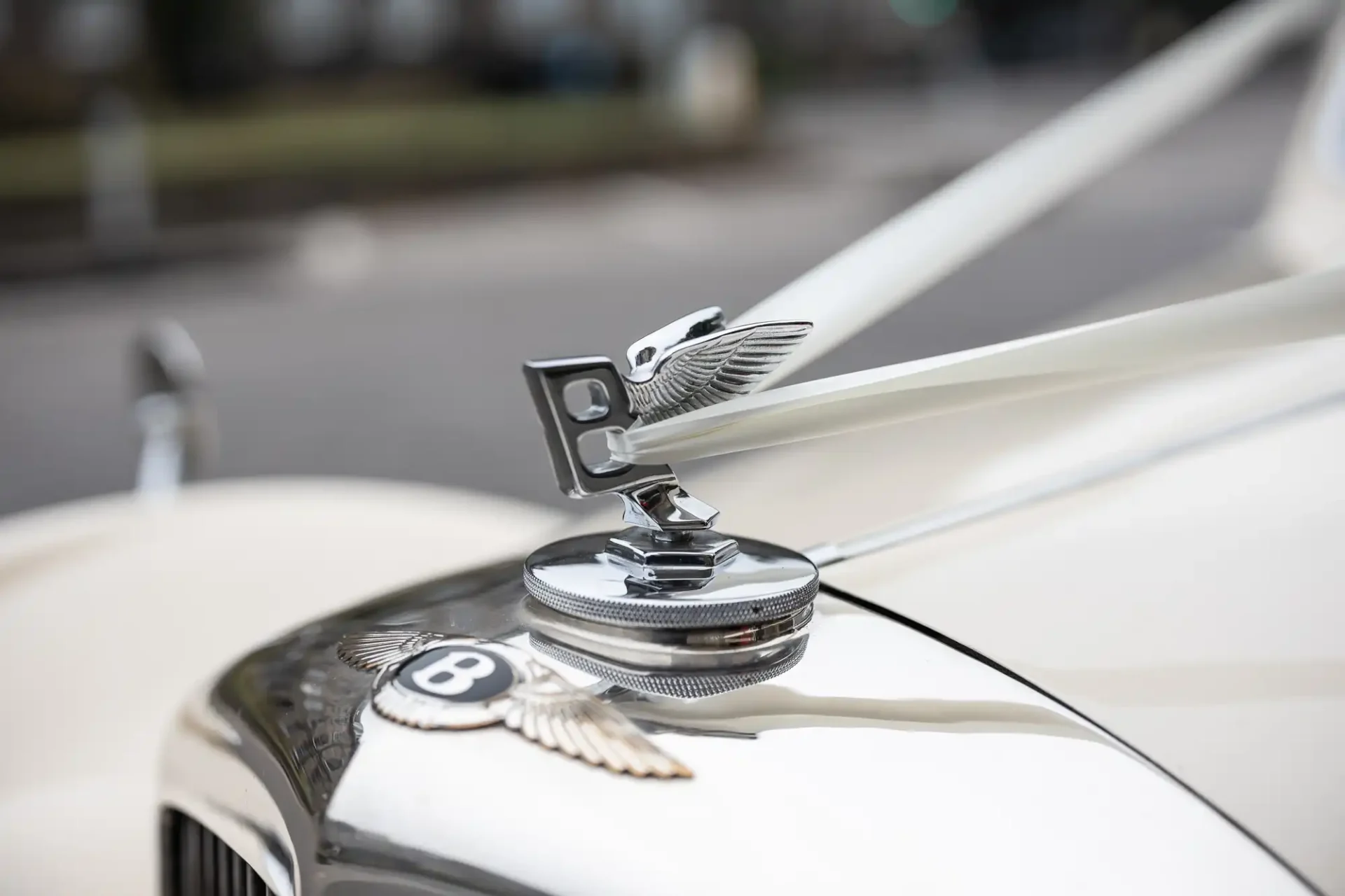 Close-up of a classic car's hood, featuring a chrome Bentley hood ornament, ribbon accents, and the Bentley logo on a cream-colored vehicle.