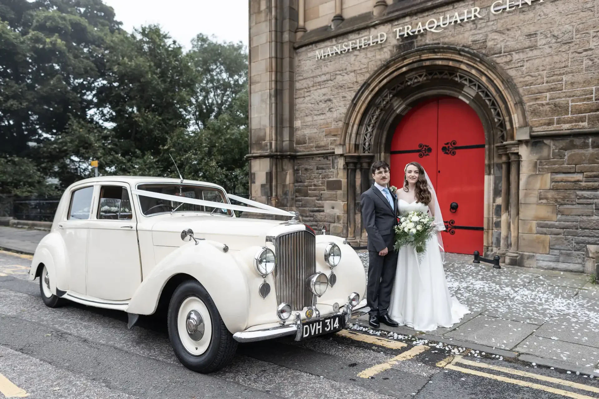 Bride and groom stand beside a vintage white car in front of a stone building with red doors.