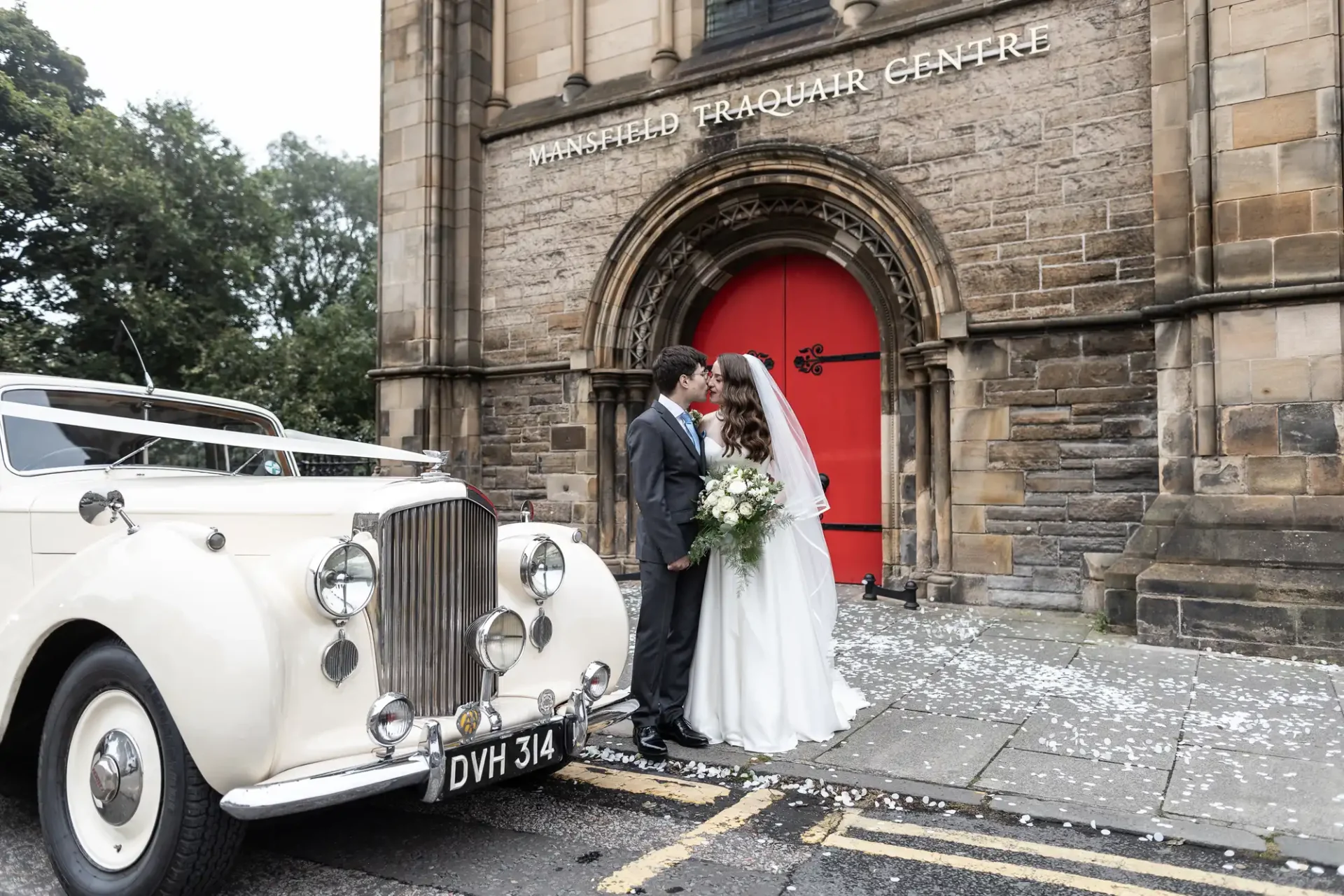 Bride and groom kiss in front of church with a red door, next to a vintage white car.