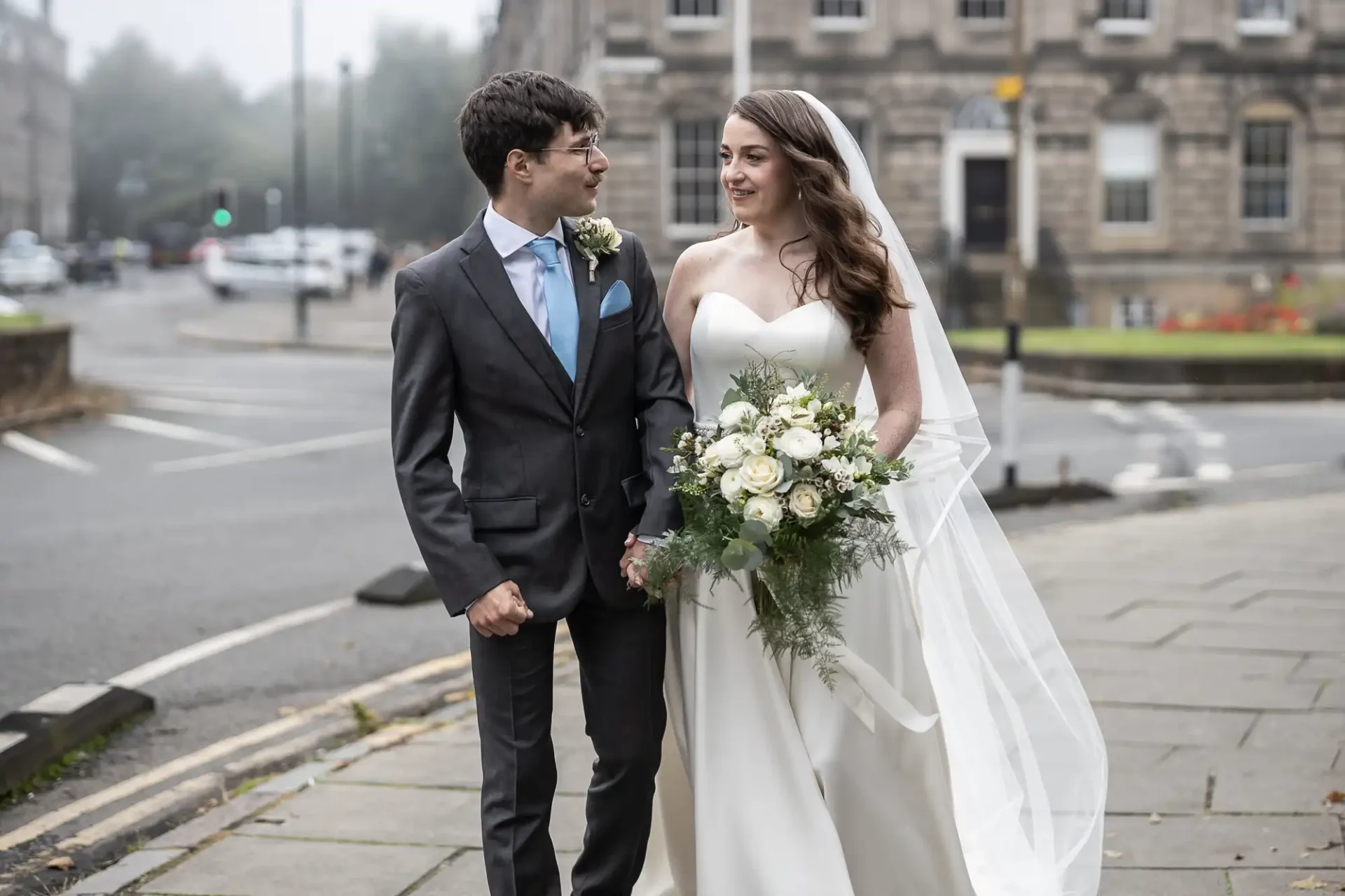 A bride and groom in formal attire walk hand in hand on an urban sidewalk, surrounded by buildings. The bride holds a bouquet of white flowers.