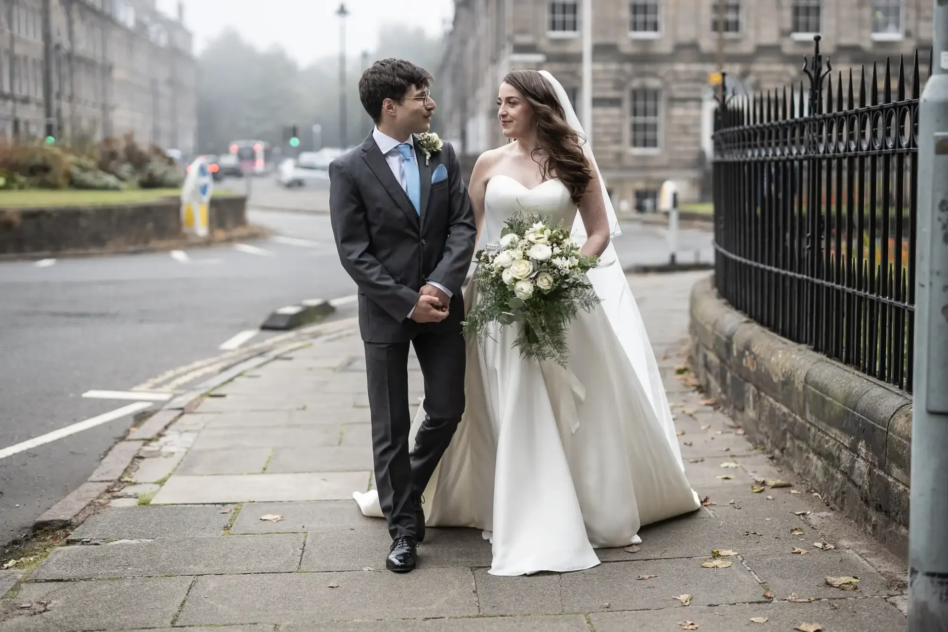 A bride in a white gown walks with a groom in a gray suit on a sidewalk. She holds a bouquet of white flowers. Buildings and a street are in the background.