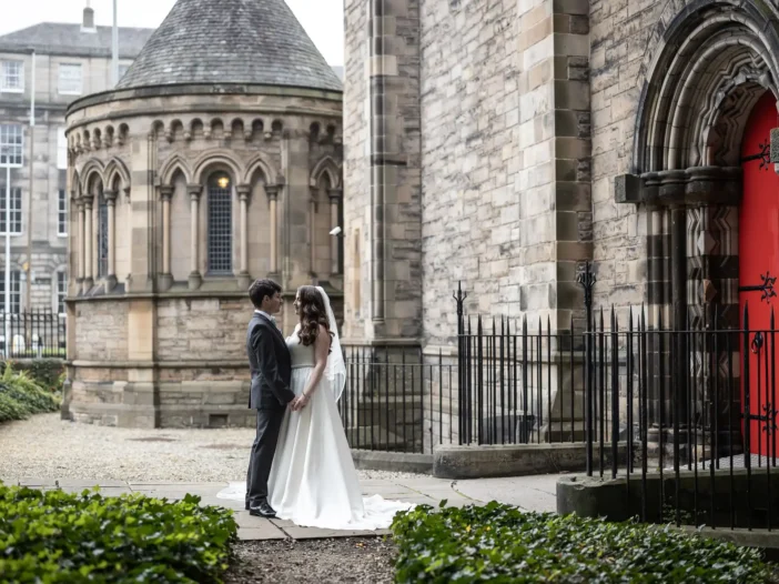 wedding photos at Mansfield Traquair: A bride and groom stand holding hands outside a historic stone building, with greenery in the foreground.