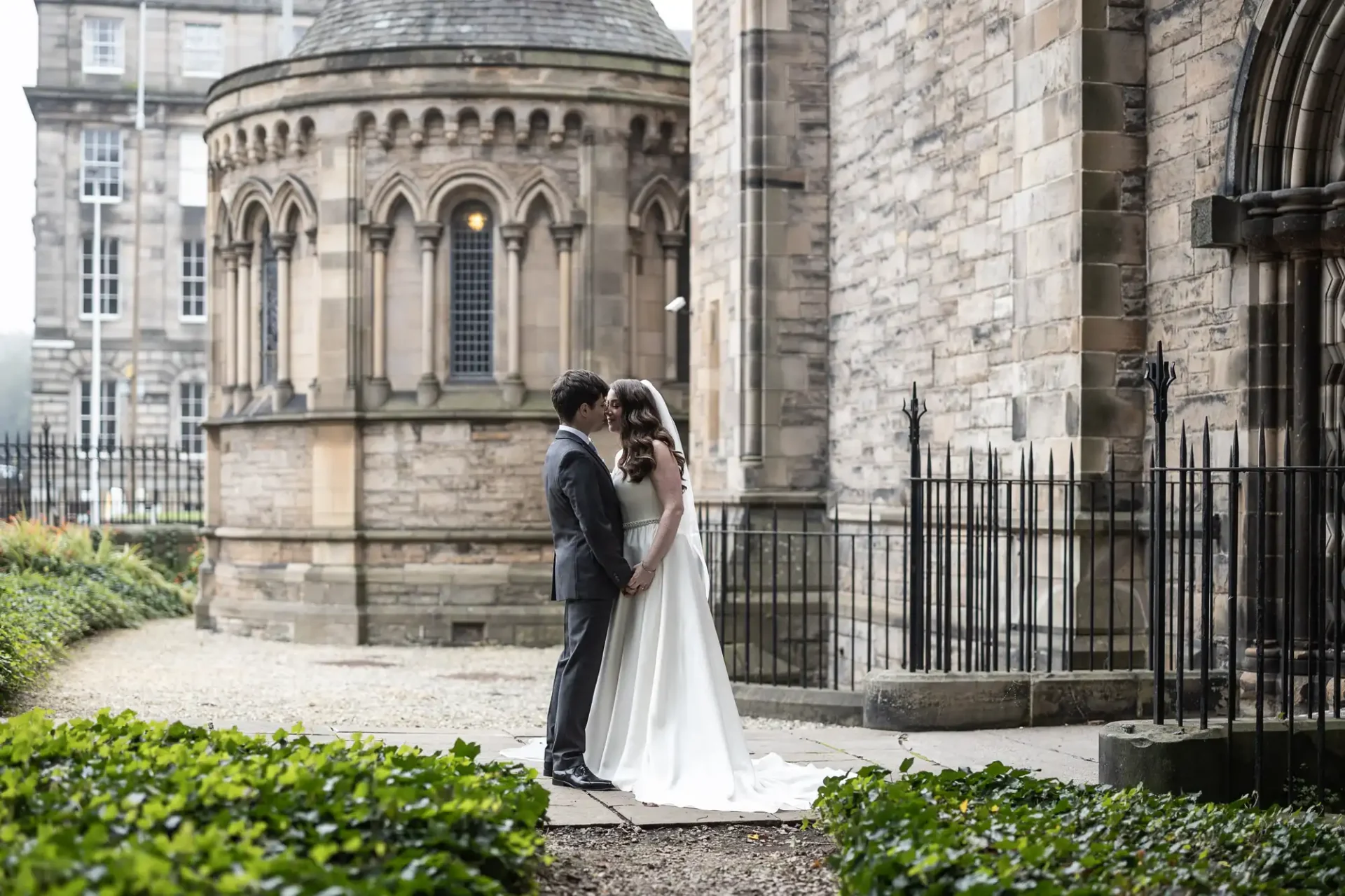 A bride and groom stand facing each other in front of an old stone building with arched windows.