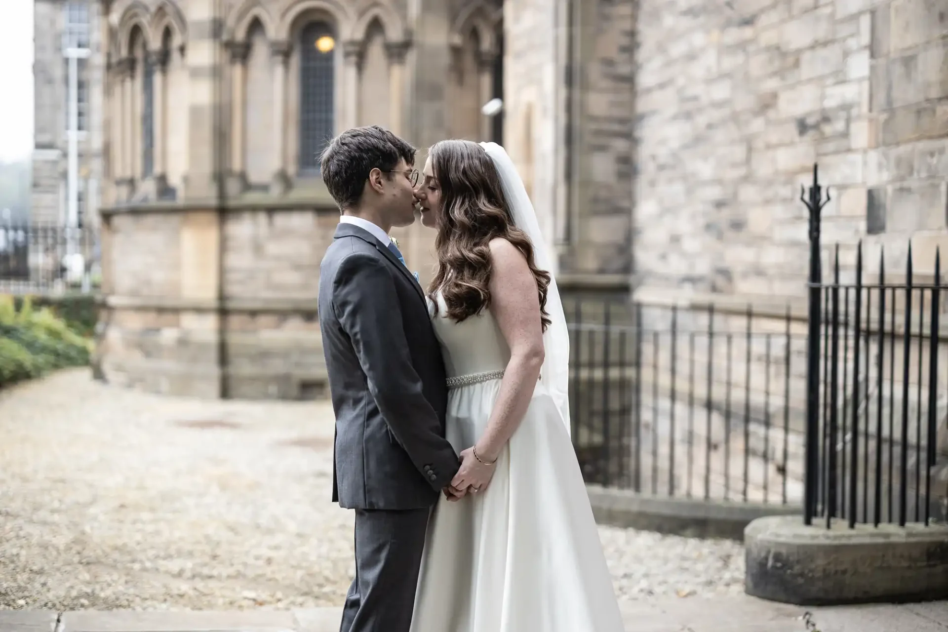 A bride and groom stand close, holding hands and touching noses, outside a stone building.