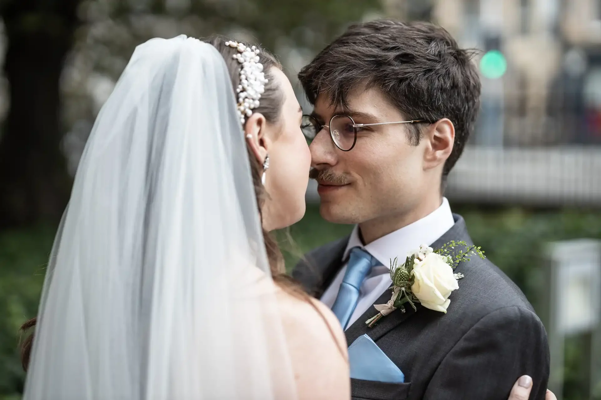 A bride and groom facing each other outdoors. The groom wears glasses and a suit with a boutonniere. The bride wears a veil and jeweled headpiece.