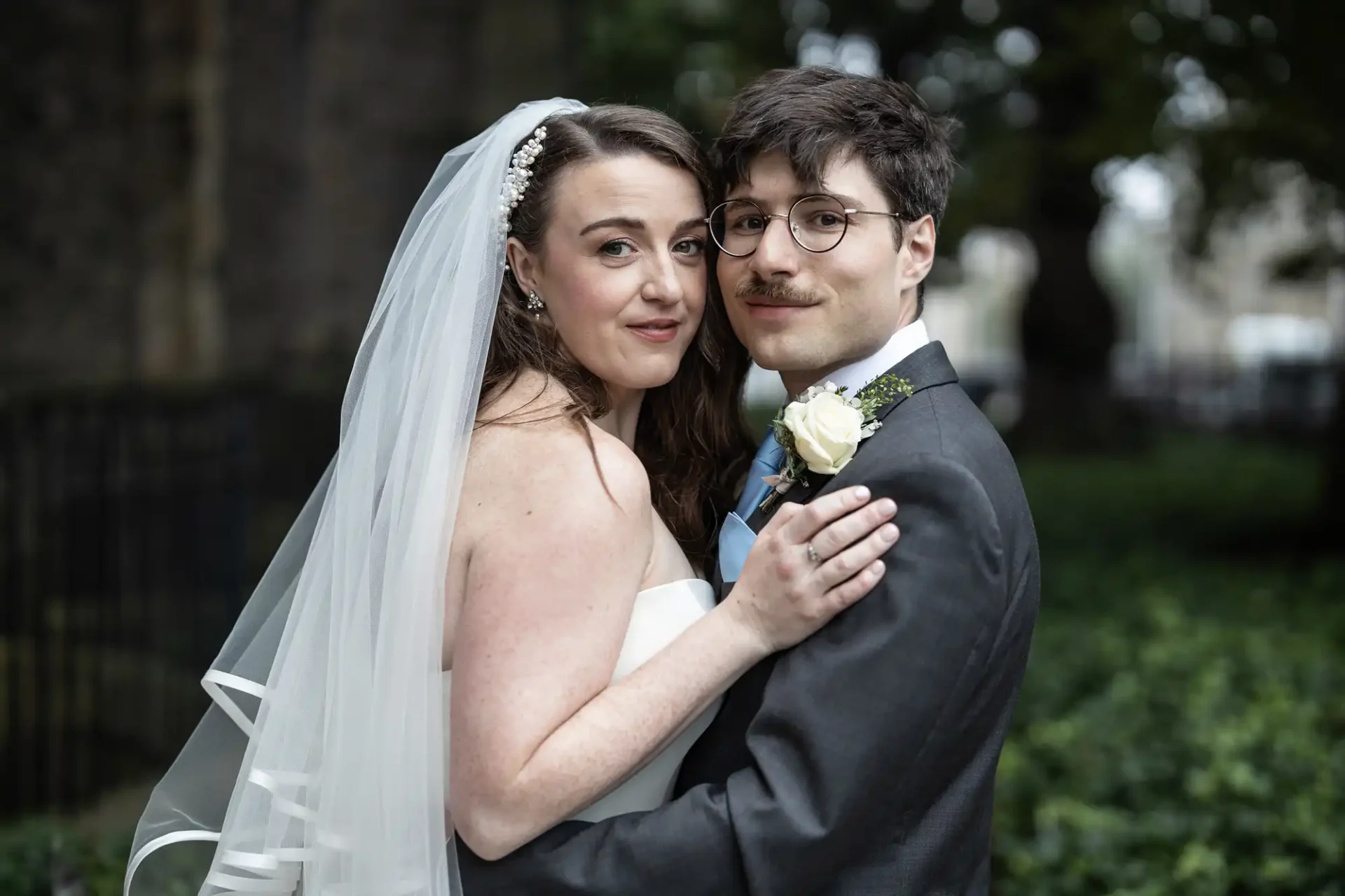 A bride in a white dress and veil embraces a groom in a suit with a boutonniere. They are outdoors, surrounded by greenery.