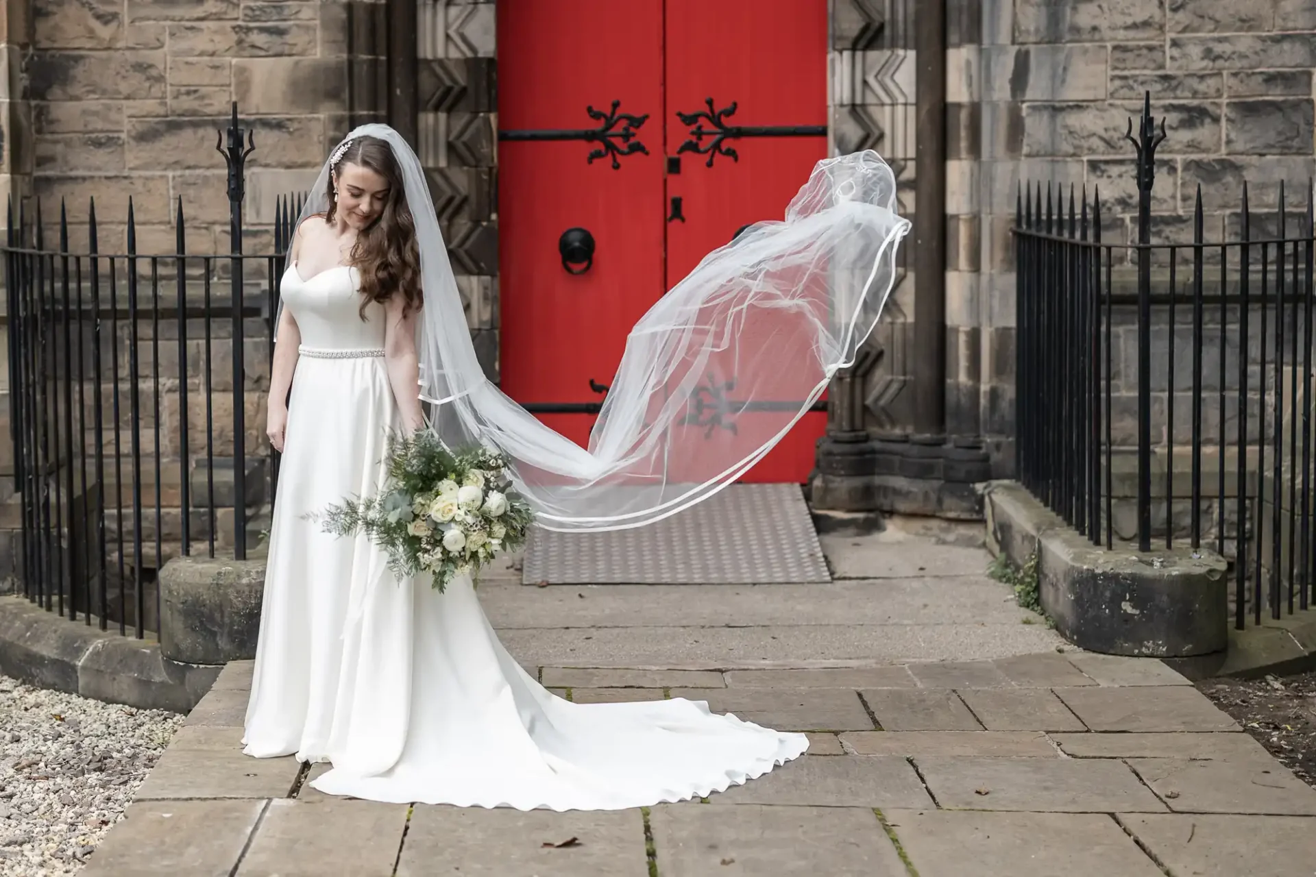 Bride in a white dress and veil holding a bouquet, standing in front of a red church door.