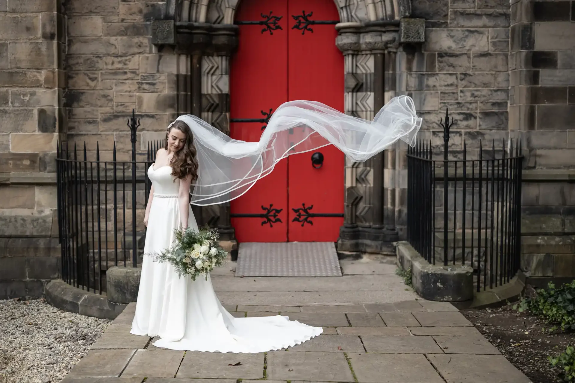 Bride in a white dress holding a bouquet stands in front of a red door, with her veil flowing behind her.