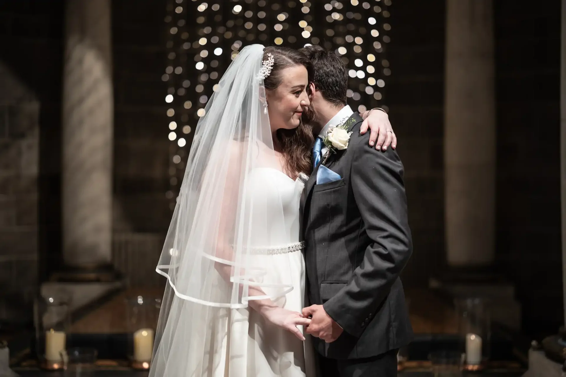 Bride and groom embrace under soft lighting, surrounded by columns and twinkling lights, holding hands, bride wearing a white dress and veil, groom in a dark suit with a floral boutonniere.