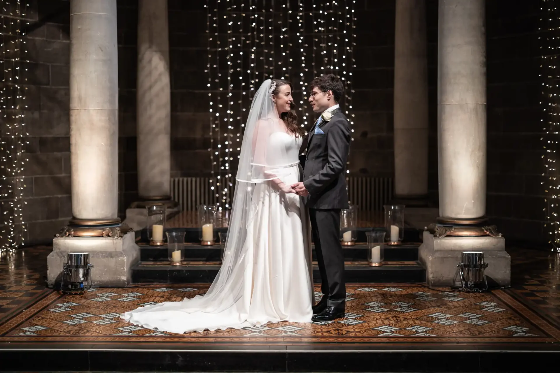 Bride and groom stand facing each other, holding hands, in a decorated venue with columns and string lights.