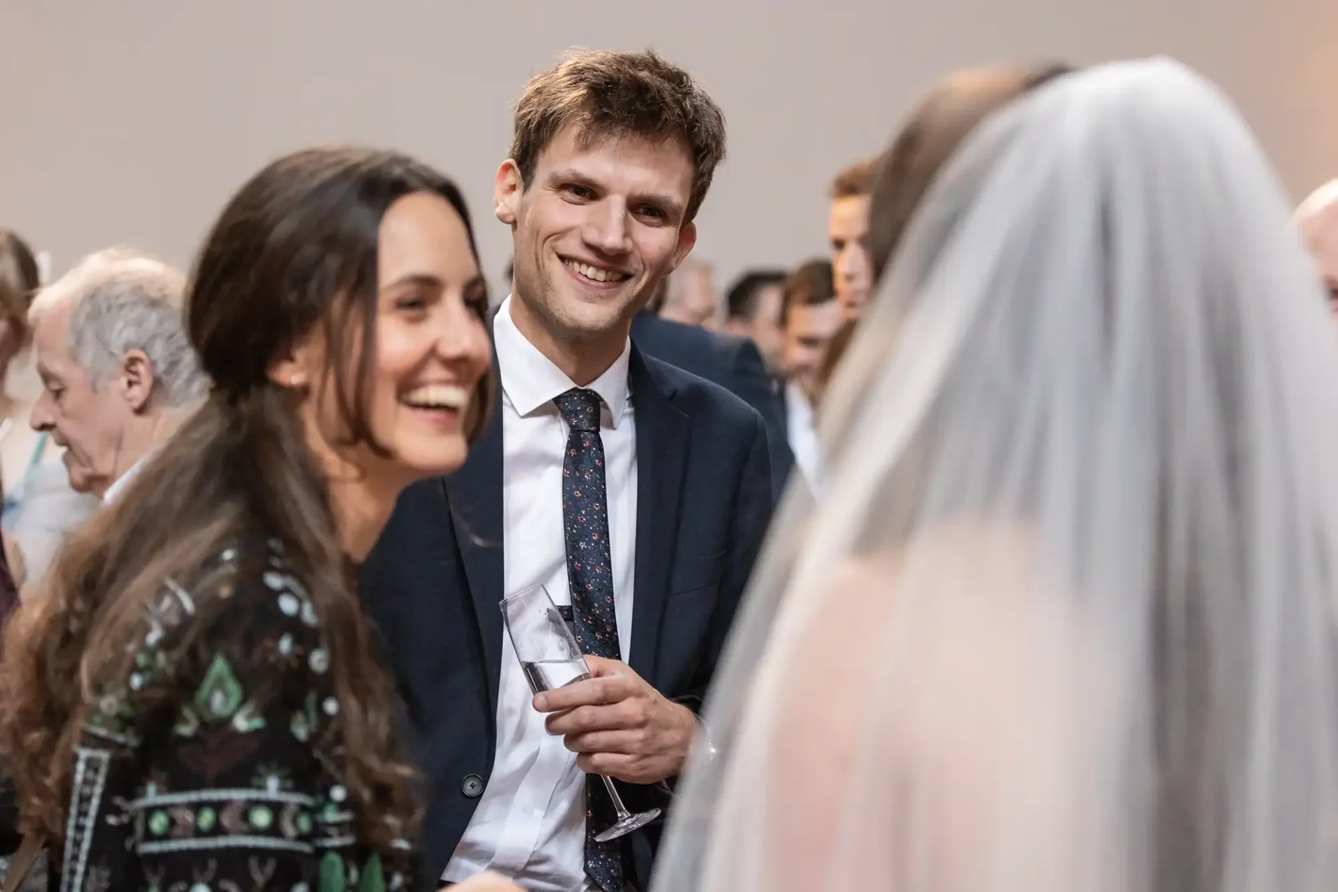 A man in a suit holding a glass talks to a woman in a wedding dress and another smiling woman at a social event.