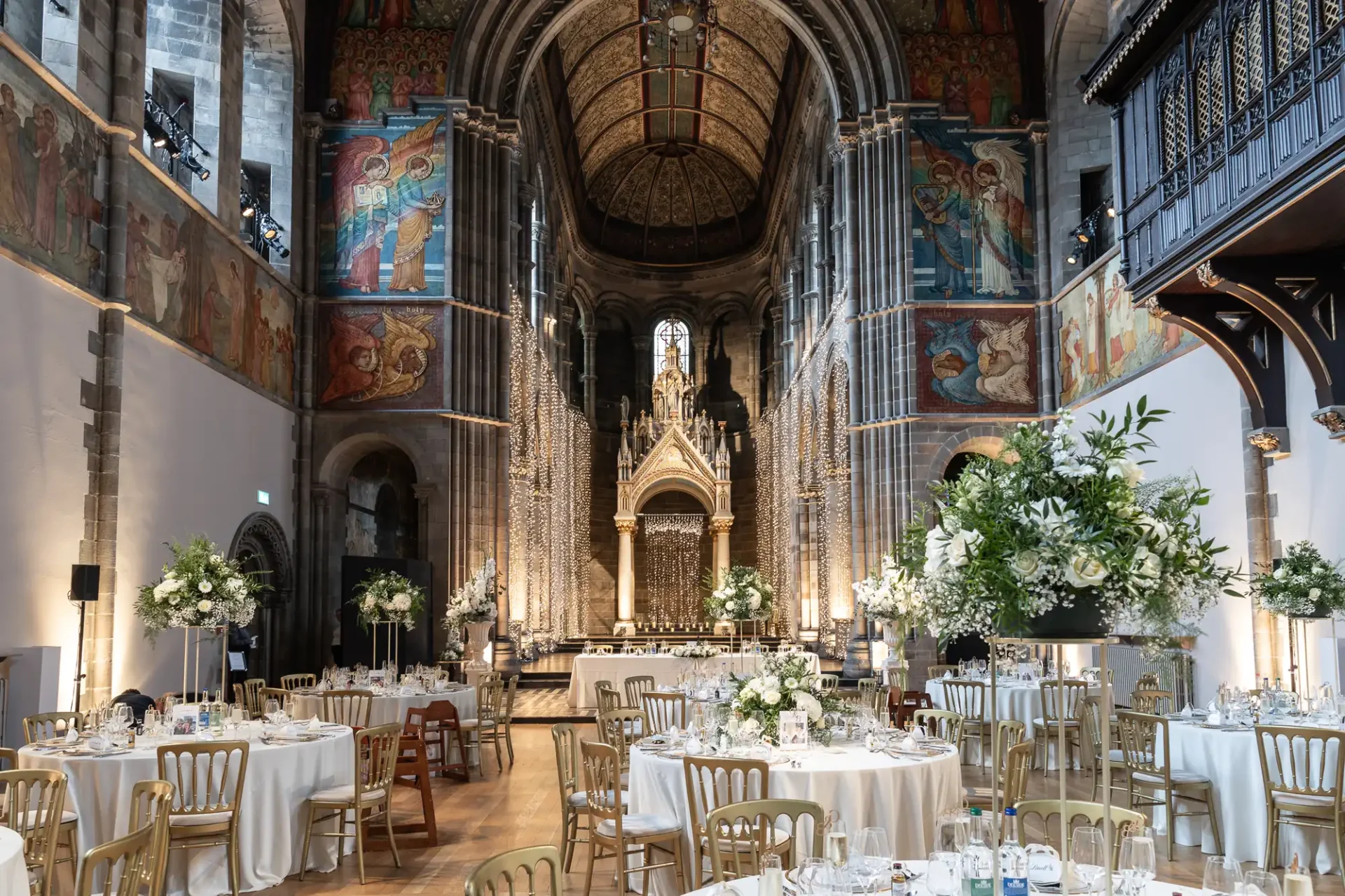 Elegant banquet setup inside a historic cathedral with ornate ceiling, hanging lights, and round tables adorned with white tablecloths and floral arrangements.