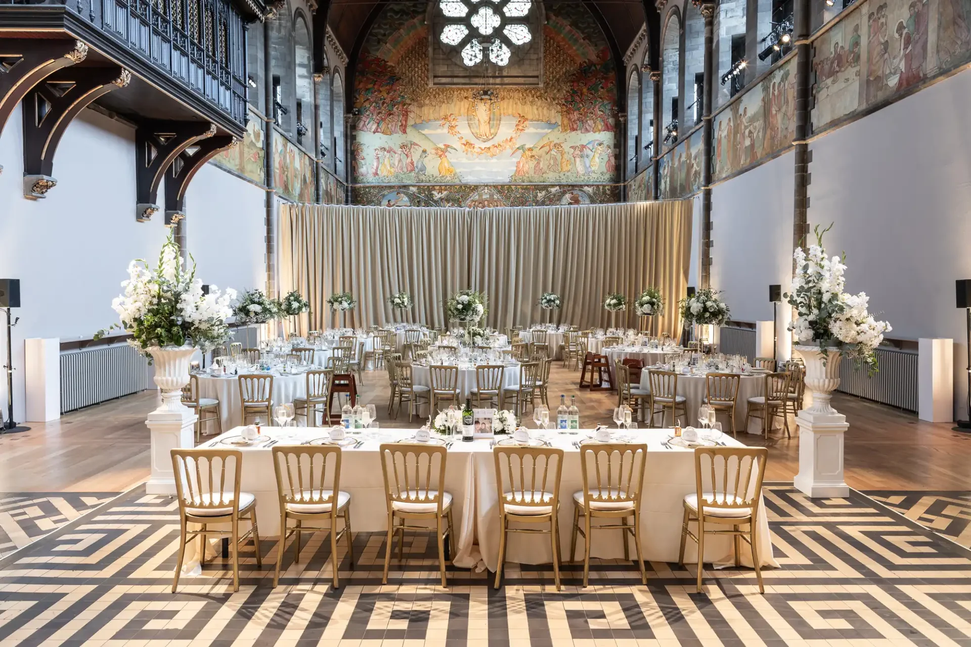 Elegant banquet hall with long tables set for a formal event, surrounded by decorative white flowers. High ceiling, patterned floor, and mural-adorned walls enhance the luxurious ambiance.