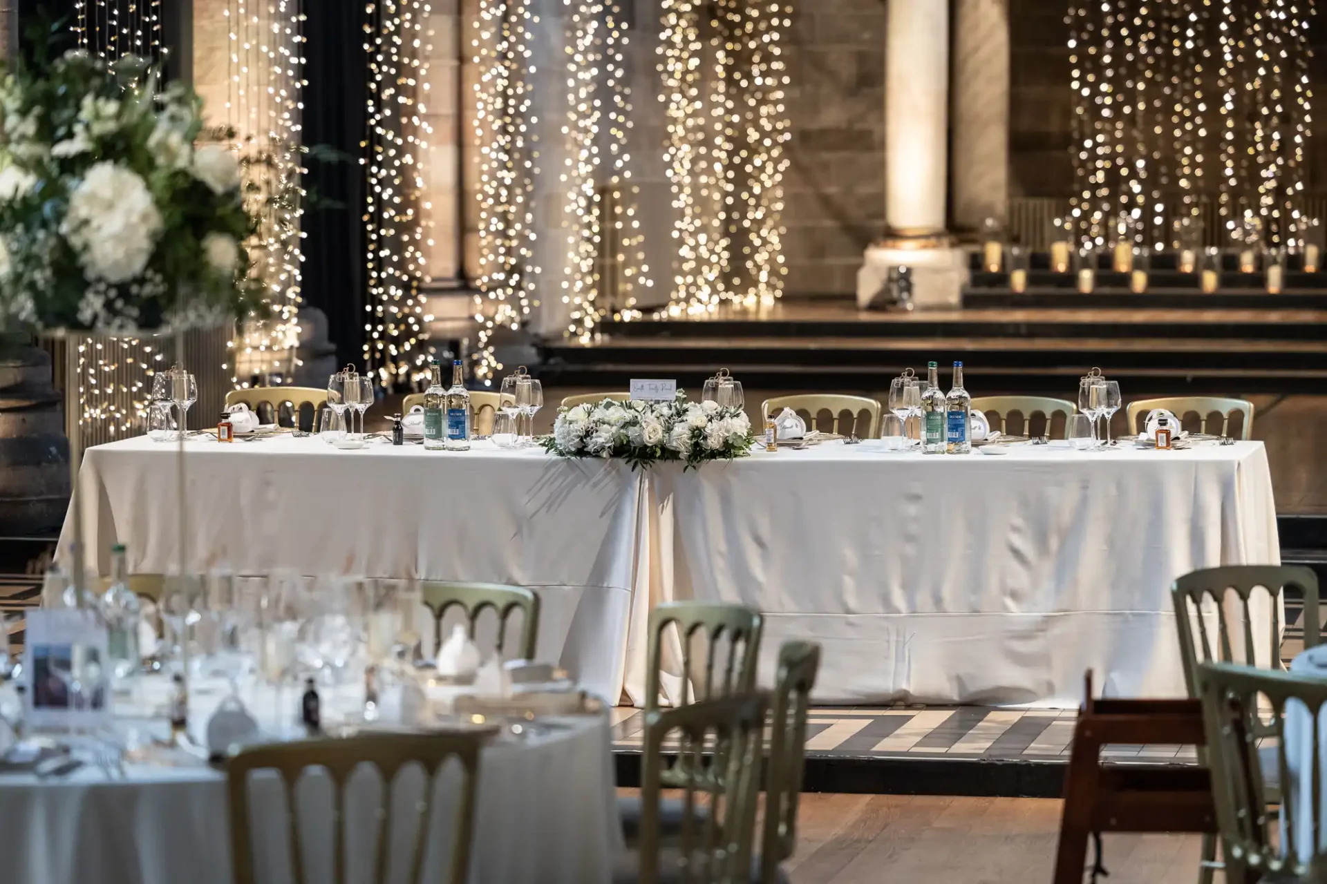 Elegant wedding reception setup with white tablecloths, floral centerpieces, and water bottles on tables. String lights and large columns decorate the background.