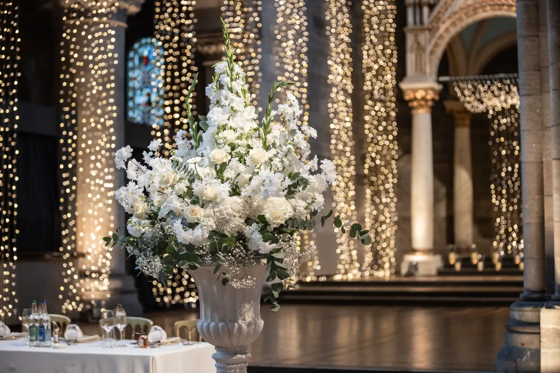 Large white floral arrangement in a decorative vase, set in an ornate hall with columns and sparkling light decorations.