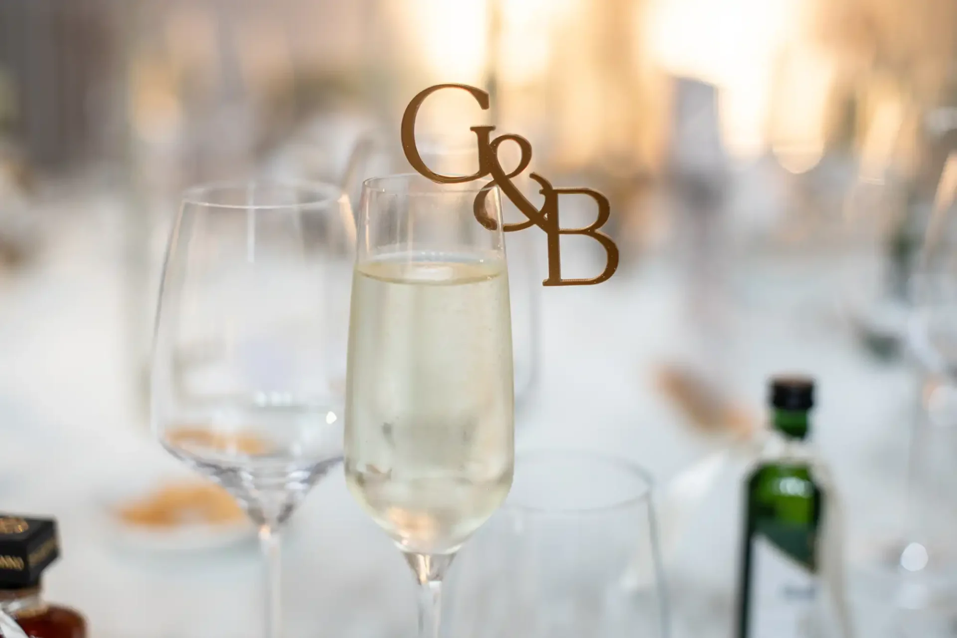 Champagne flute with initials "G&B" on a pick, surrounded by empty glassware and small bottles on a blurred background.