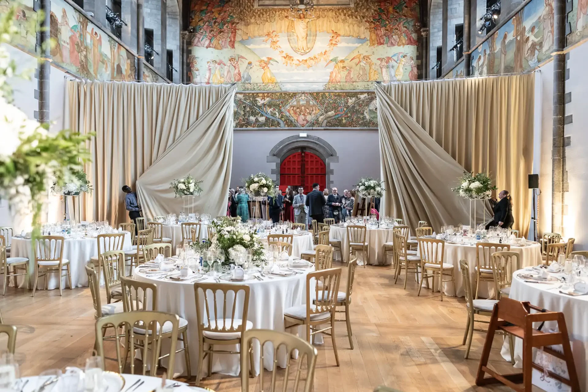 A grand banquet hall with decorated round tables, flower arrangements, and draped curtains. Guests are gathering near the entrance beneath a large mural on the wall.