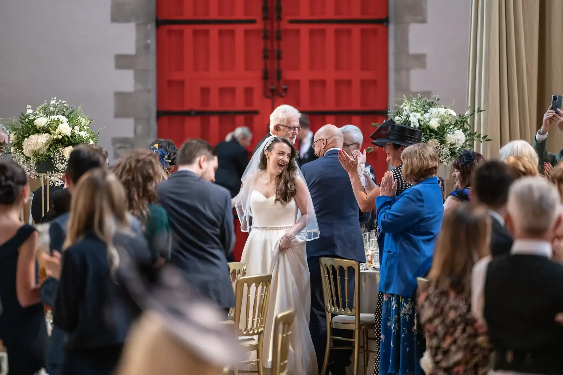 A bride in a white dress walks down an aisle, smiling amid applauding guests. She's near tables with floral arrangements, and a large red door is visible in the background.