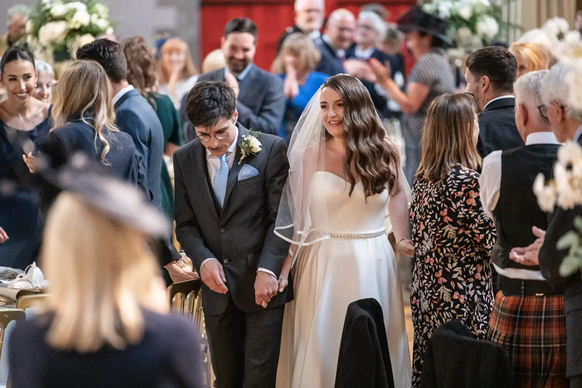 A bride and groom walk down the aisle, smiling and holding hands. They are surrounded by standing, applauding guests in formal attire at a wedding ceremony.