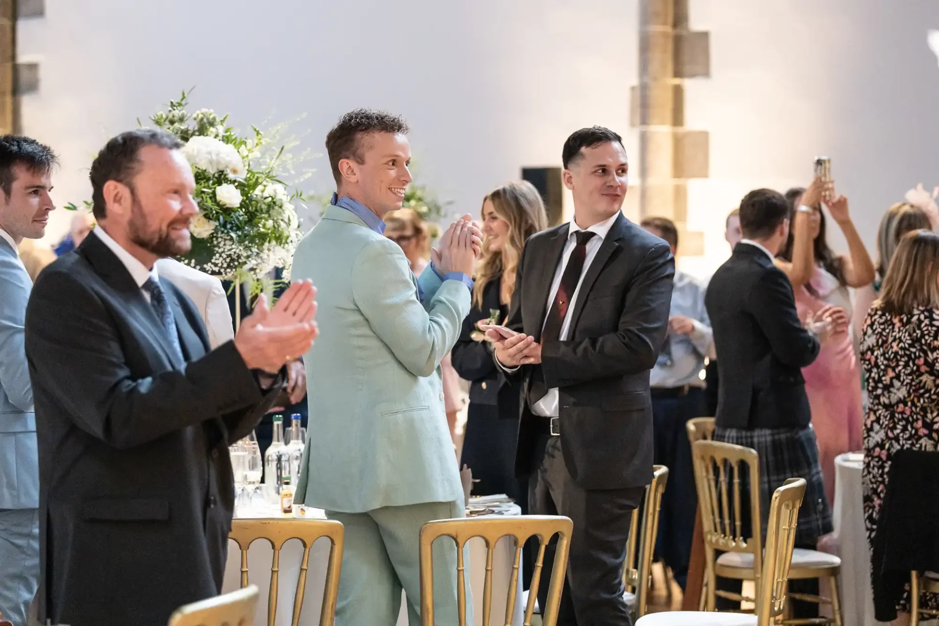 Group of people in formal attire stand and applaud in a brightly lit room with round tables and floral centerpieces.