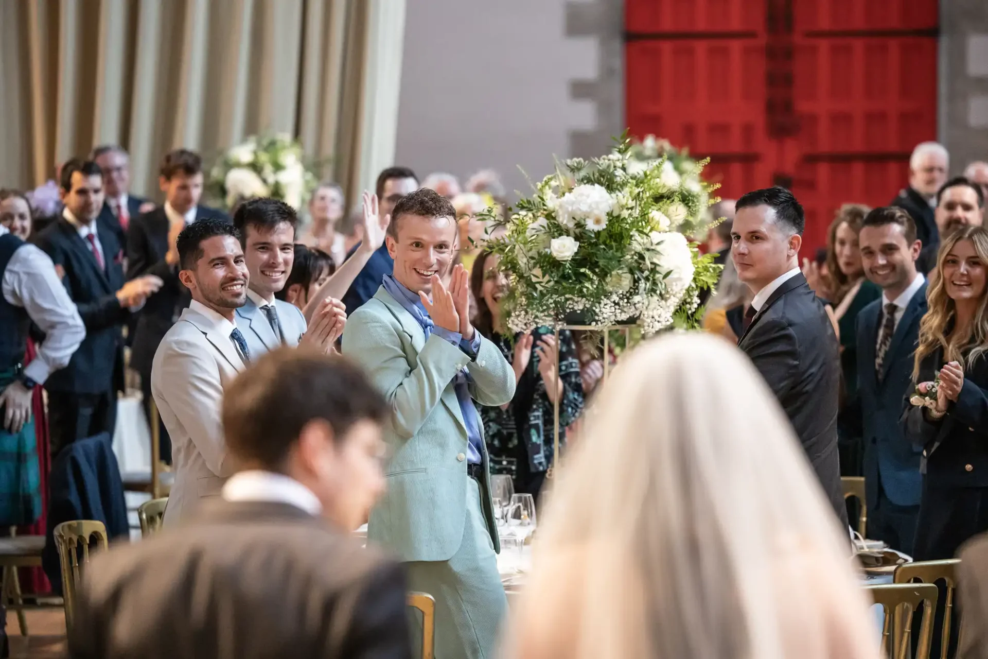 Guests stand and applaud as a bride enters a wedding reception, with a focus on a man in a light blue suit clapping.