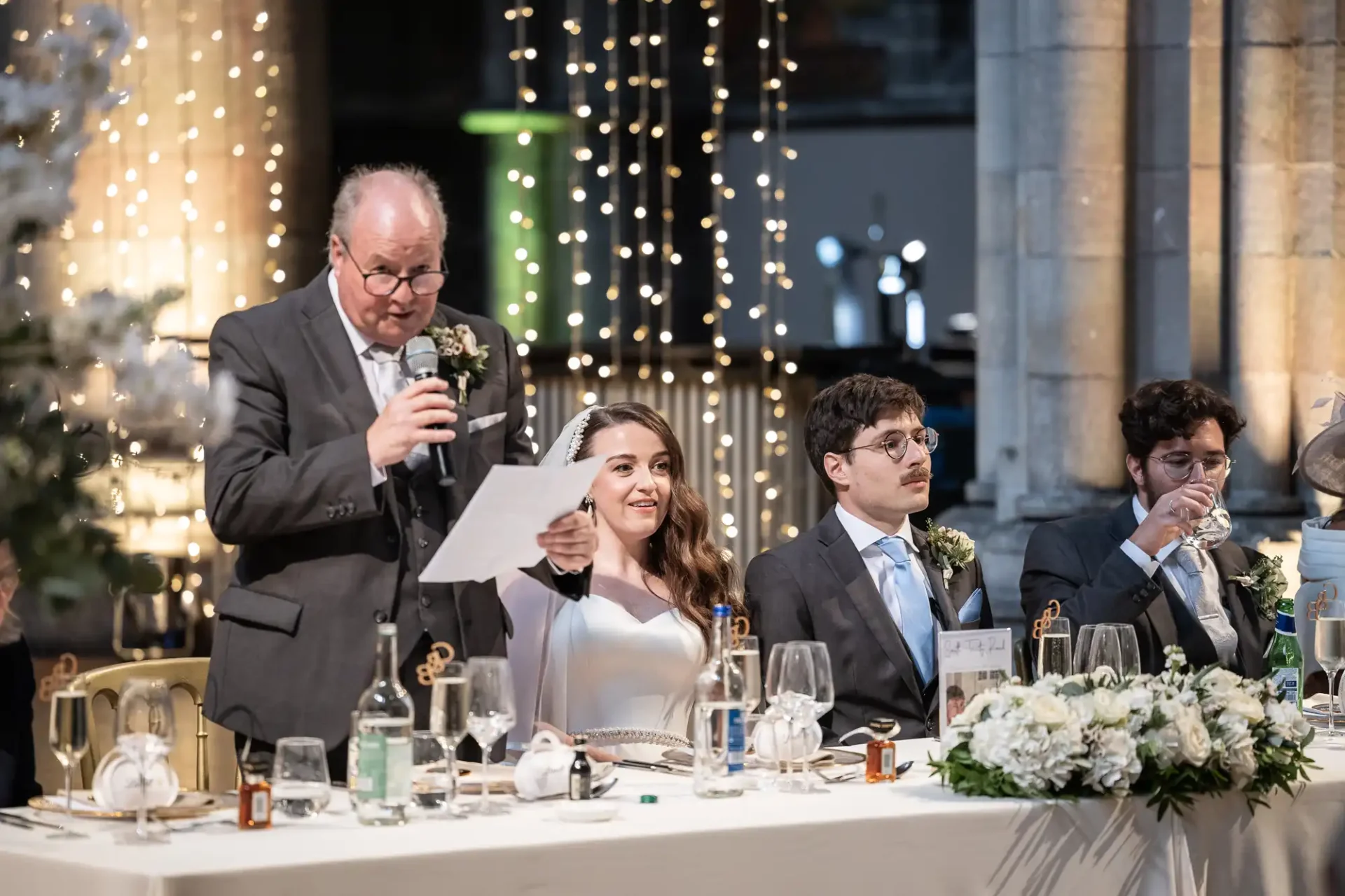 A person delivers a speech at a wedding reception. The bride, seated with two others, smiles. Tables are set with glasses, bottles, and floral arrangements. String lights hang in the background.