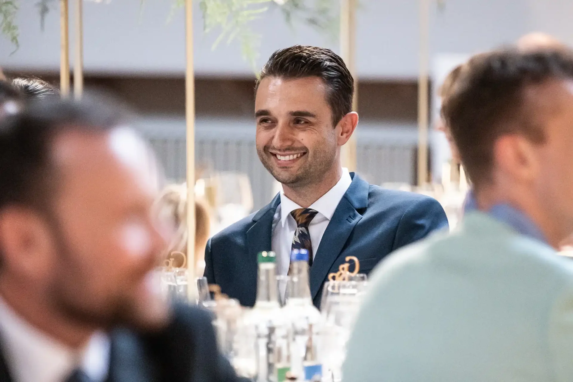 A man in a suit smiling and sitting at a table during an event, surrounded by bottles and glasses, with other people nearby.