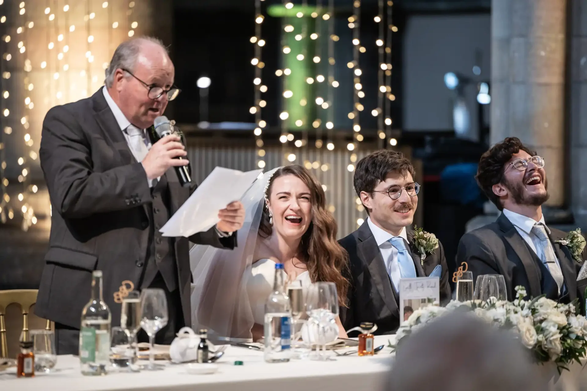 A man in a suit speaks into a microphone next to a seated couple and another man at a wedding reception. They are all laughing, with a string of lights in the background.