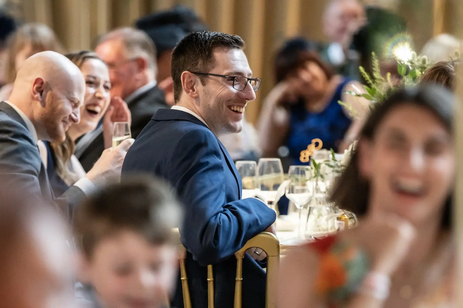 Guests seated at a formal event, smiling and laughing, with a focus on a man in glasses and a suit.