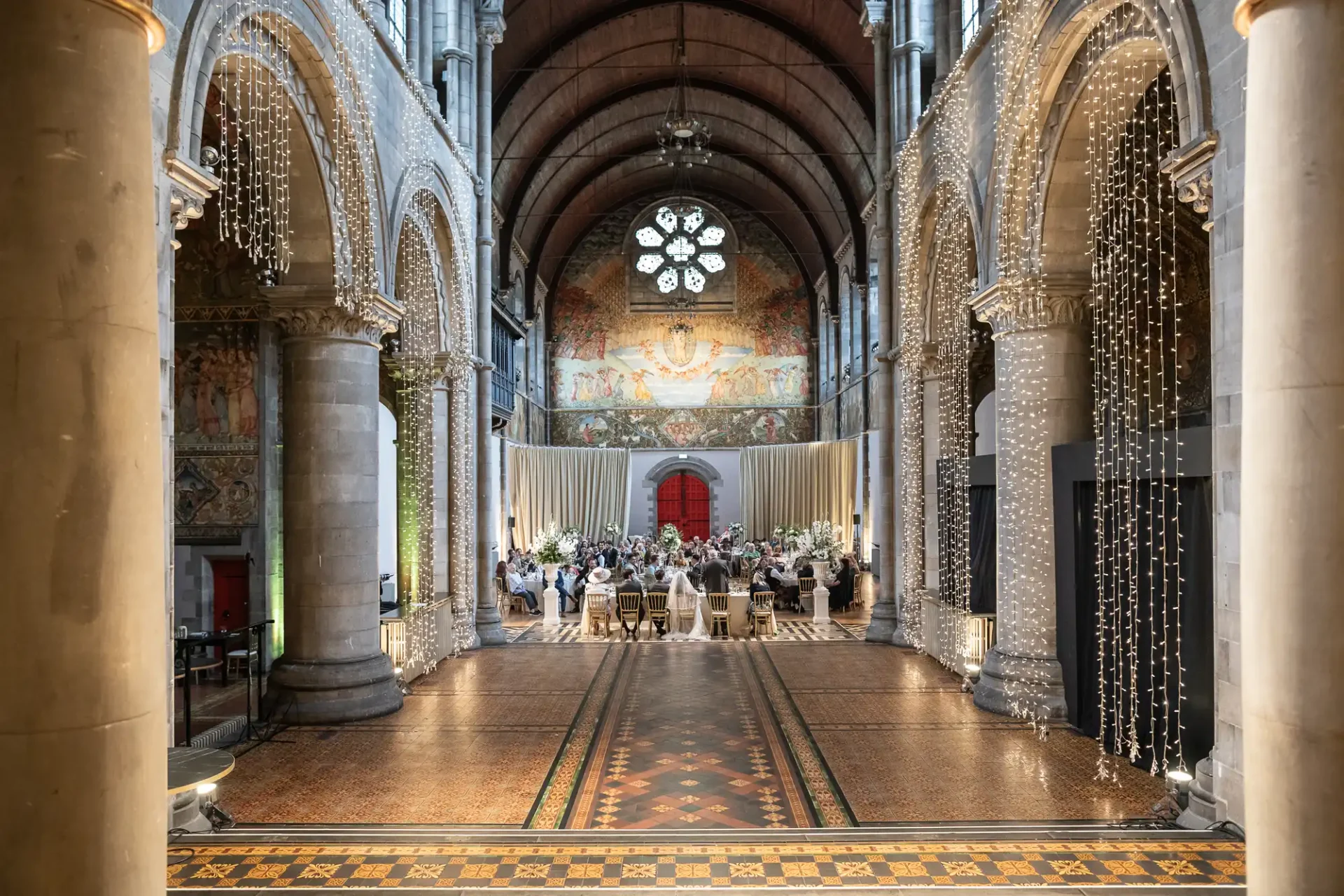 A grand hall with vaulted ceiling and tall arches, featuring a dining event setup with round tables. A large circular stained glass window is visible at the far end.