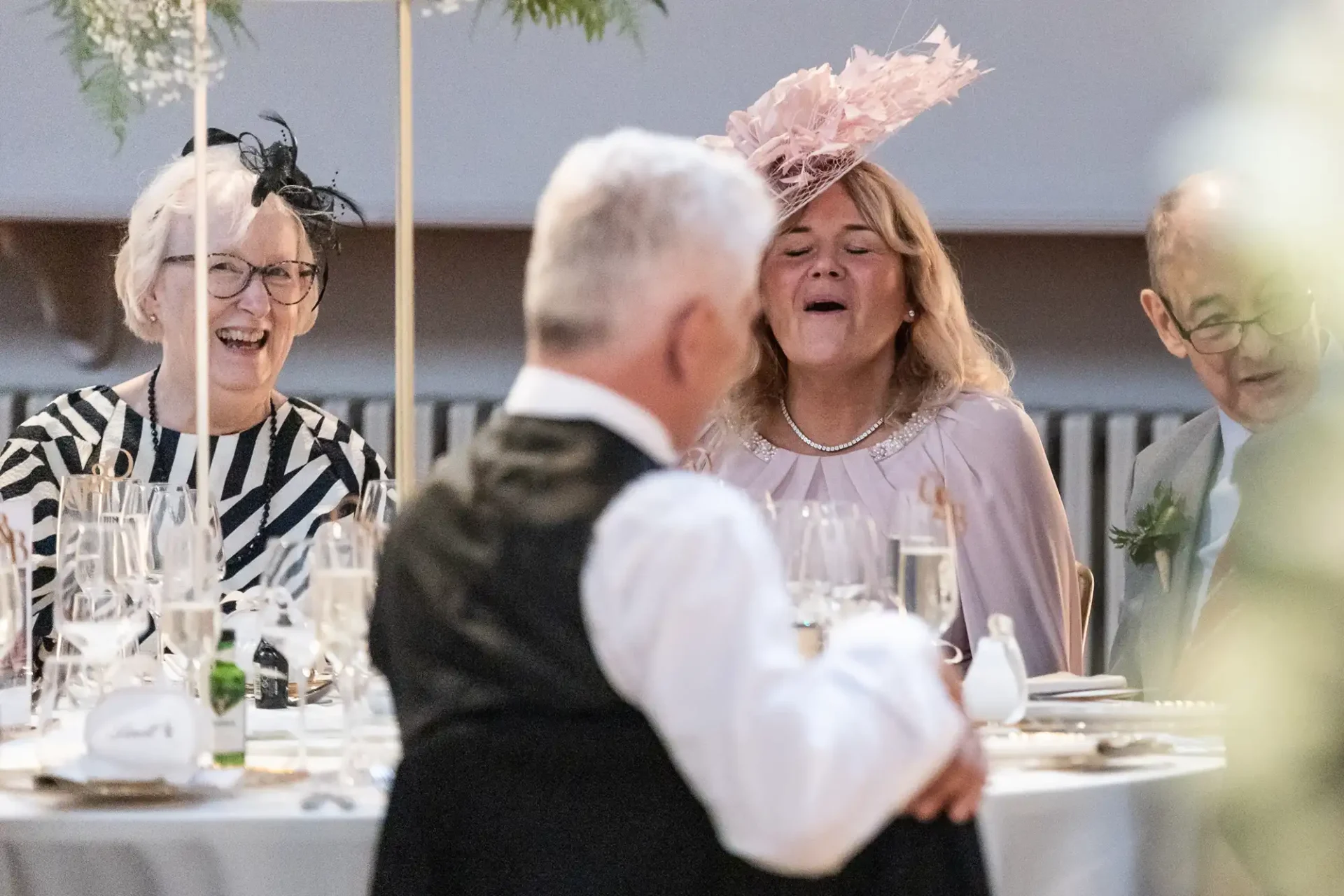 Three people are seated at a table dressed elegantly, with one woman laughing. They are surrounded by glasses and plates, suggesting they are at a formal event or gathering.