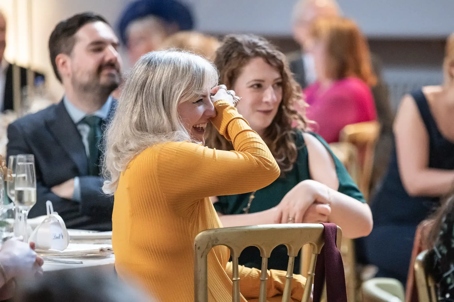 A woman in a mustard sweater, smiling and covering her mouth, sits at a table with others at an event. Glasses and plates are visible in the background.