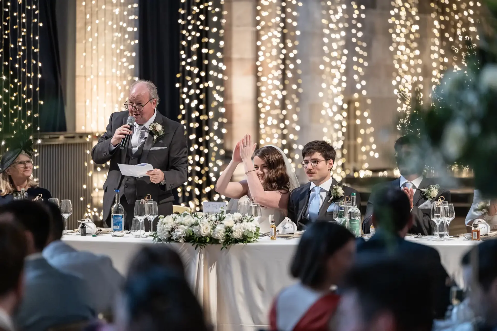 A person speaks into a microphone at a wedding reception. A woman seated at the table appears to be clapping or raising her hand. A man sits beside her. String lights decorate the background.