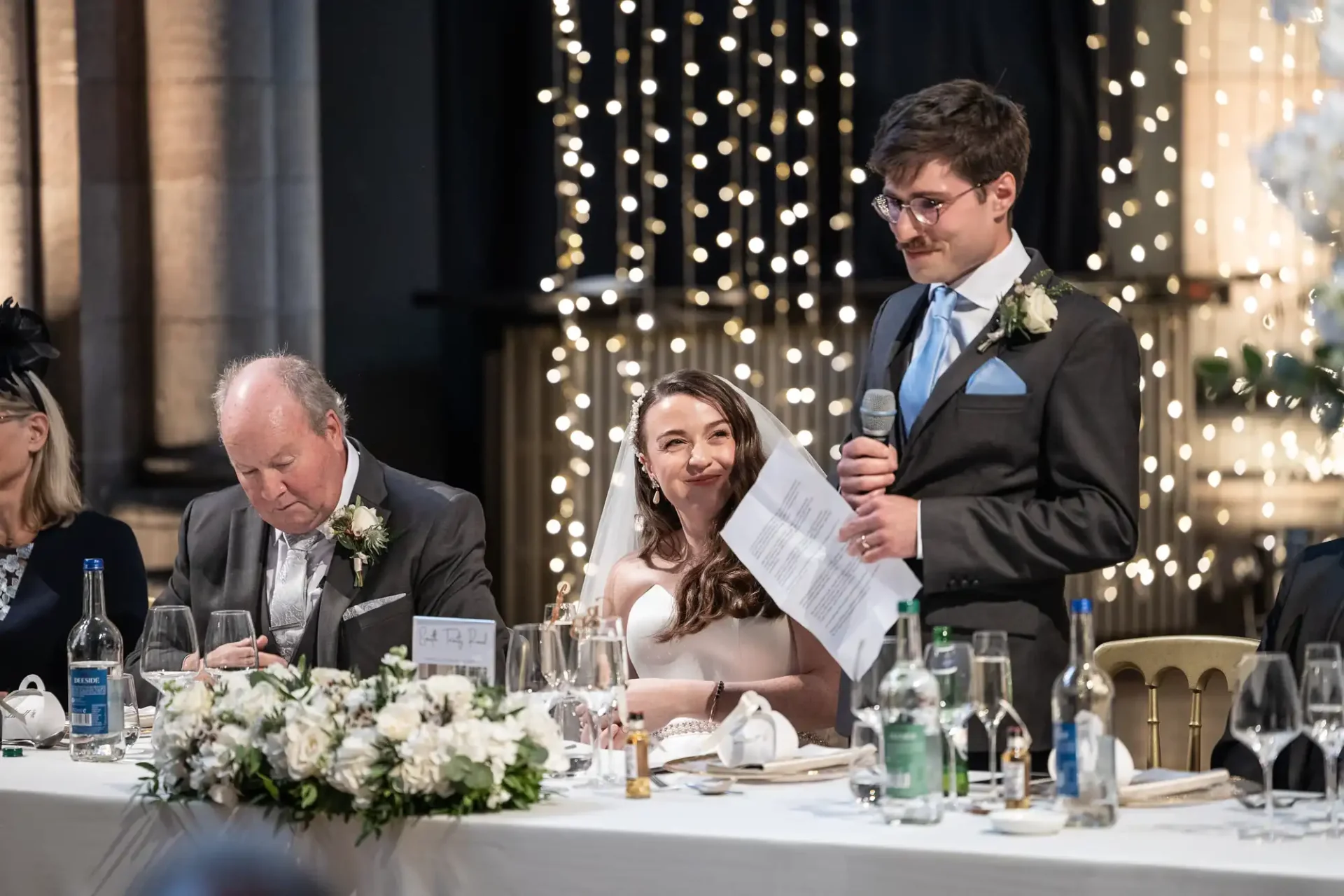 A man in a suit gives a speech at a wedding reception. A woman in a white dress sits smiling beside him at a decorated table, with string lights in the background.