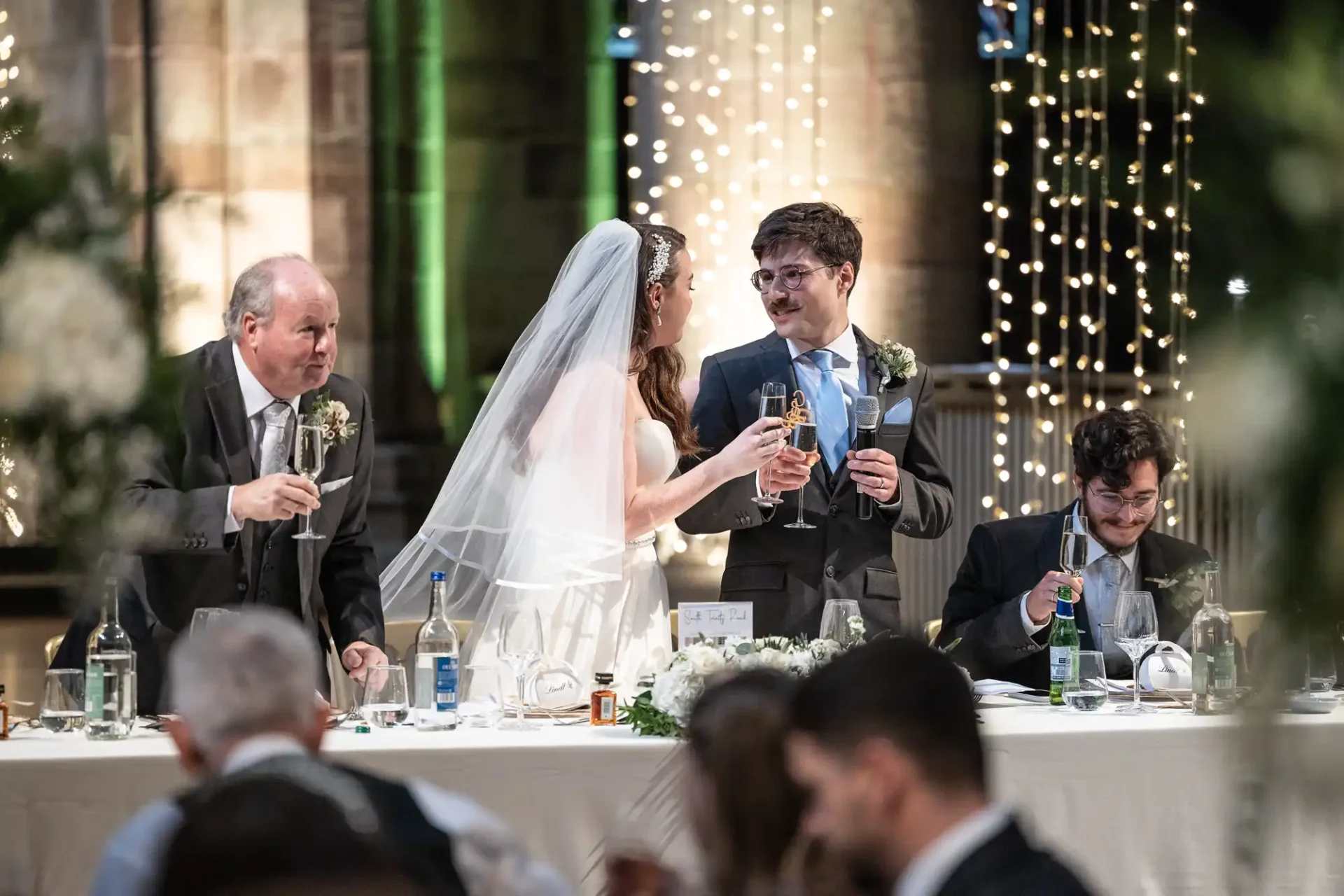 Bride and groom share a toast with guests at a wedding reception. A man on the left and another on the right also hold glasses. The table is decorated with flowers and bottles.