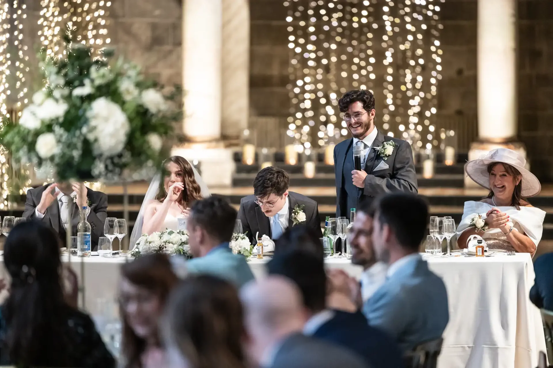 A man gives a speech at a wedding reception. Guests sit at a long, decorated table with flowers and lights in the background.