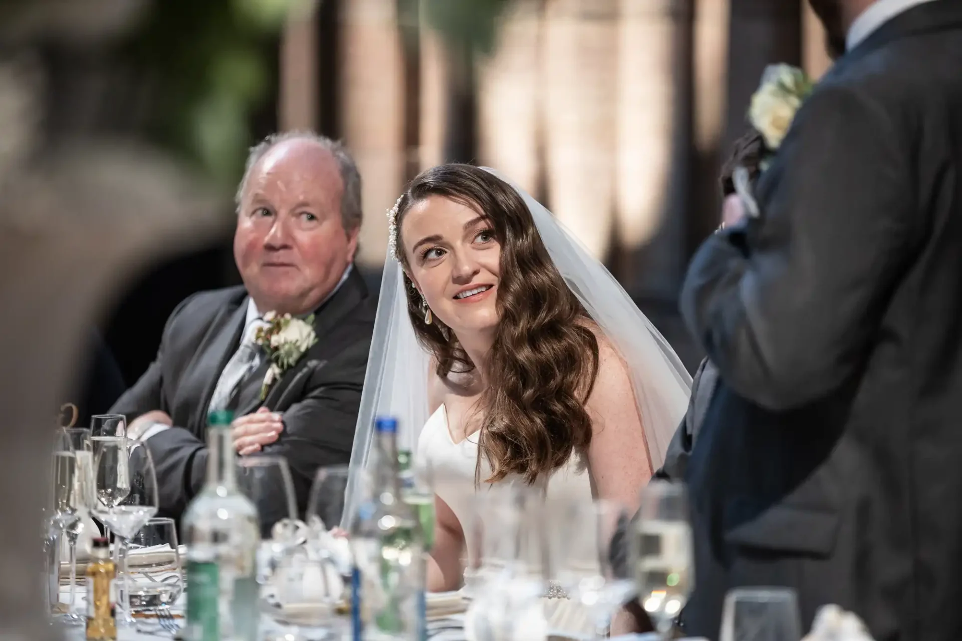 Bride in a white dress and veil sits at a table, looking up, while an older man in a suit looks on.