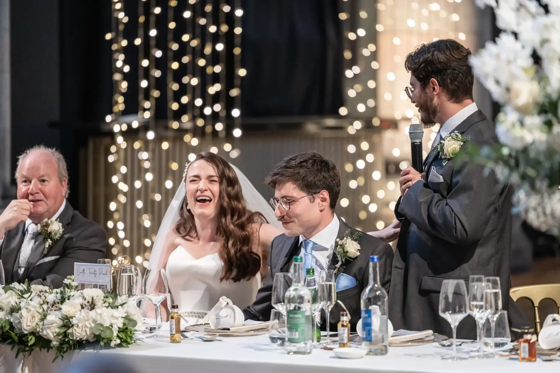 A bride and groom sit at a wedding reception table adorned with flowers. The groom is looking down, while a man in a suit stands, speaking into a microphone. Fairy lights hang in the background.