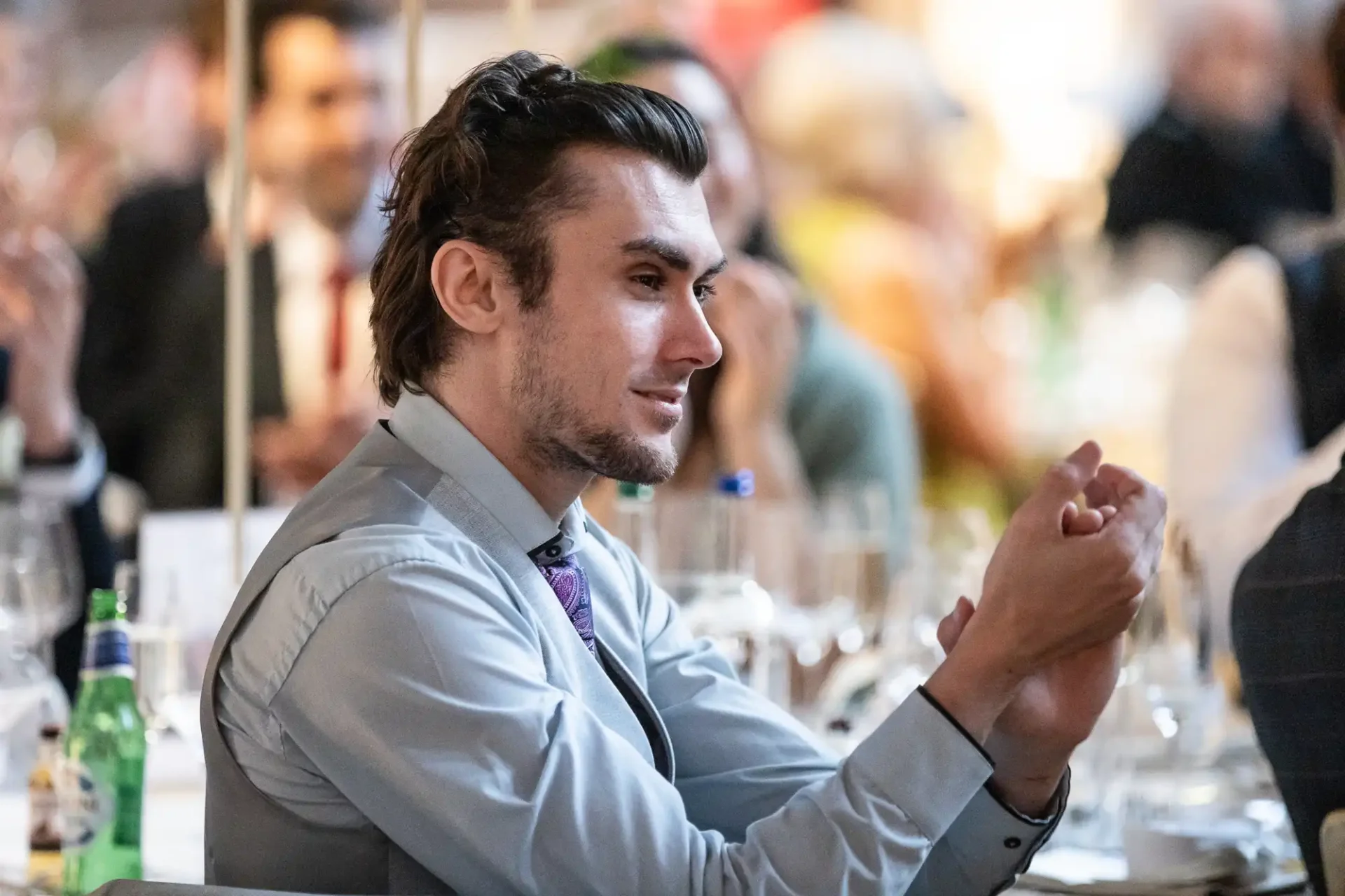 Person in a gray shirt claps while seated at an event, surrounded by tables and other attendees.