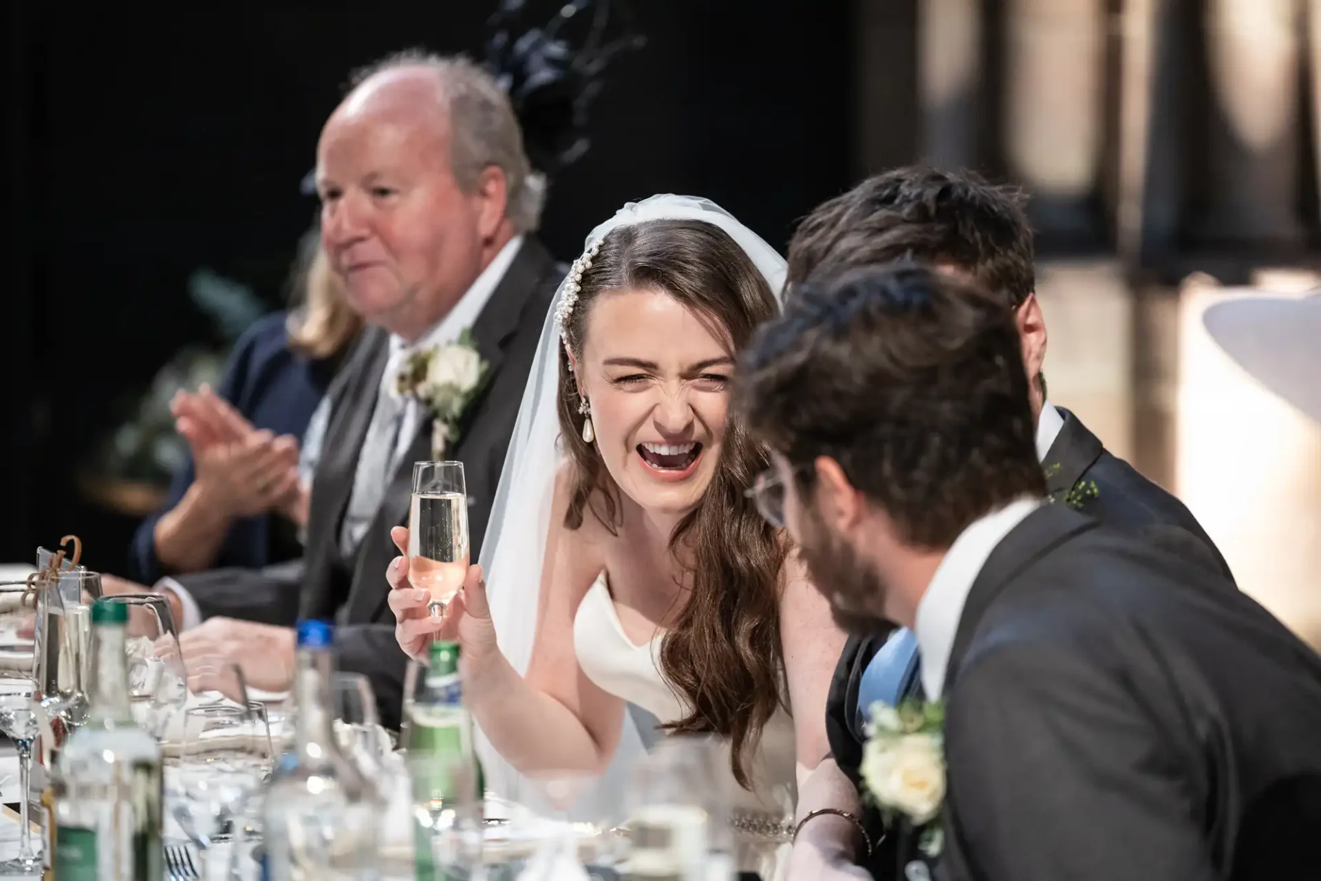 Bride laughing with a glass in hand at a dining table during a wedding reception, surrounded by guests in formal attire.