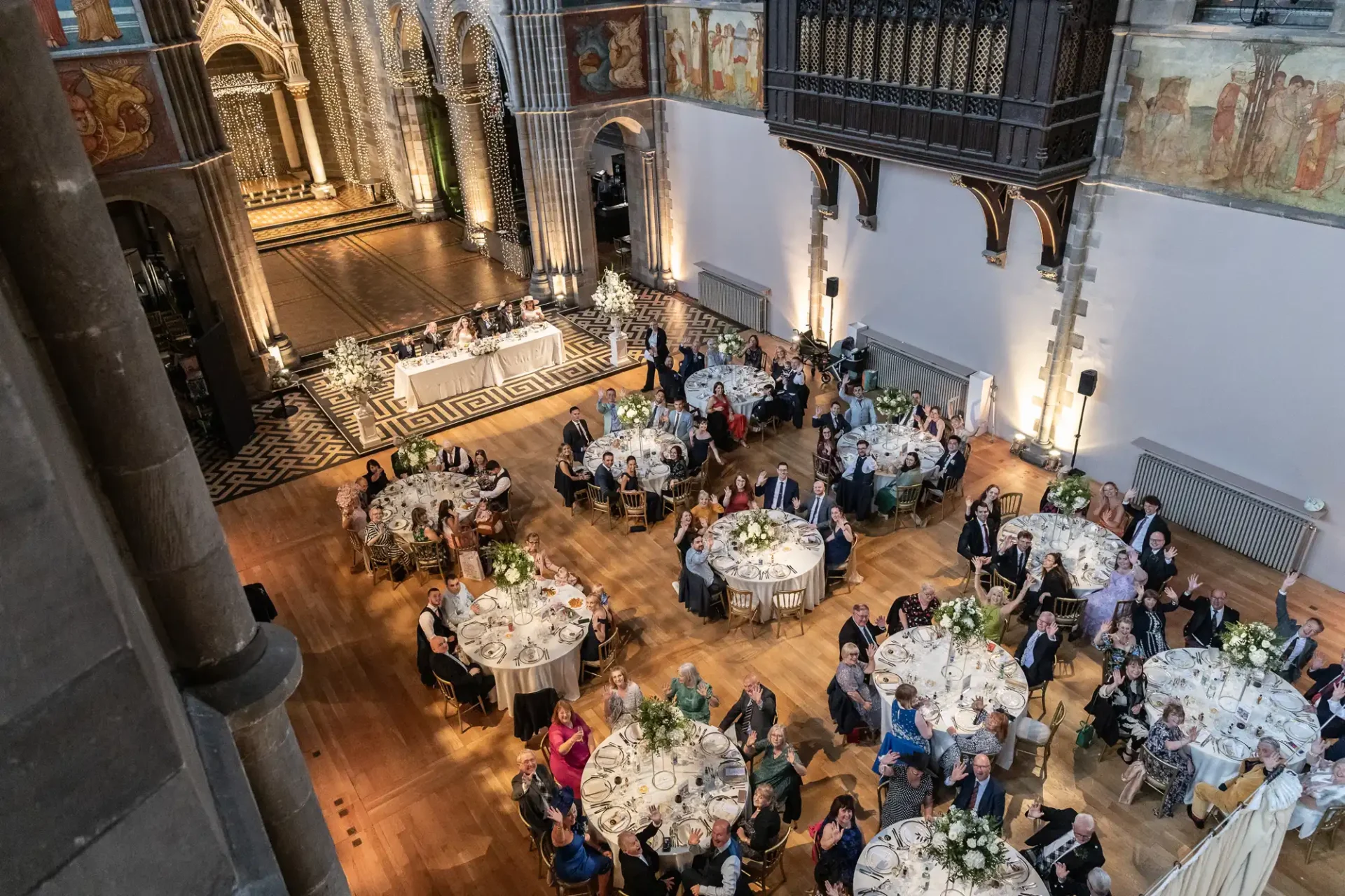 Aerial view of a formal banquet in a grand hall with round tables filled with seated guests. The hall features ornate architectural details and decorative lighting.