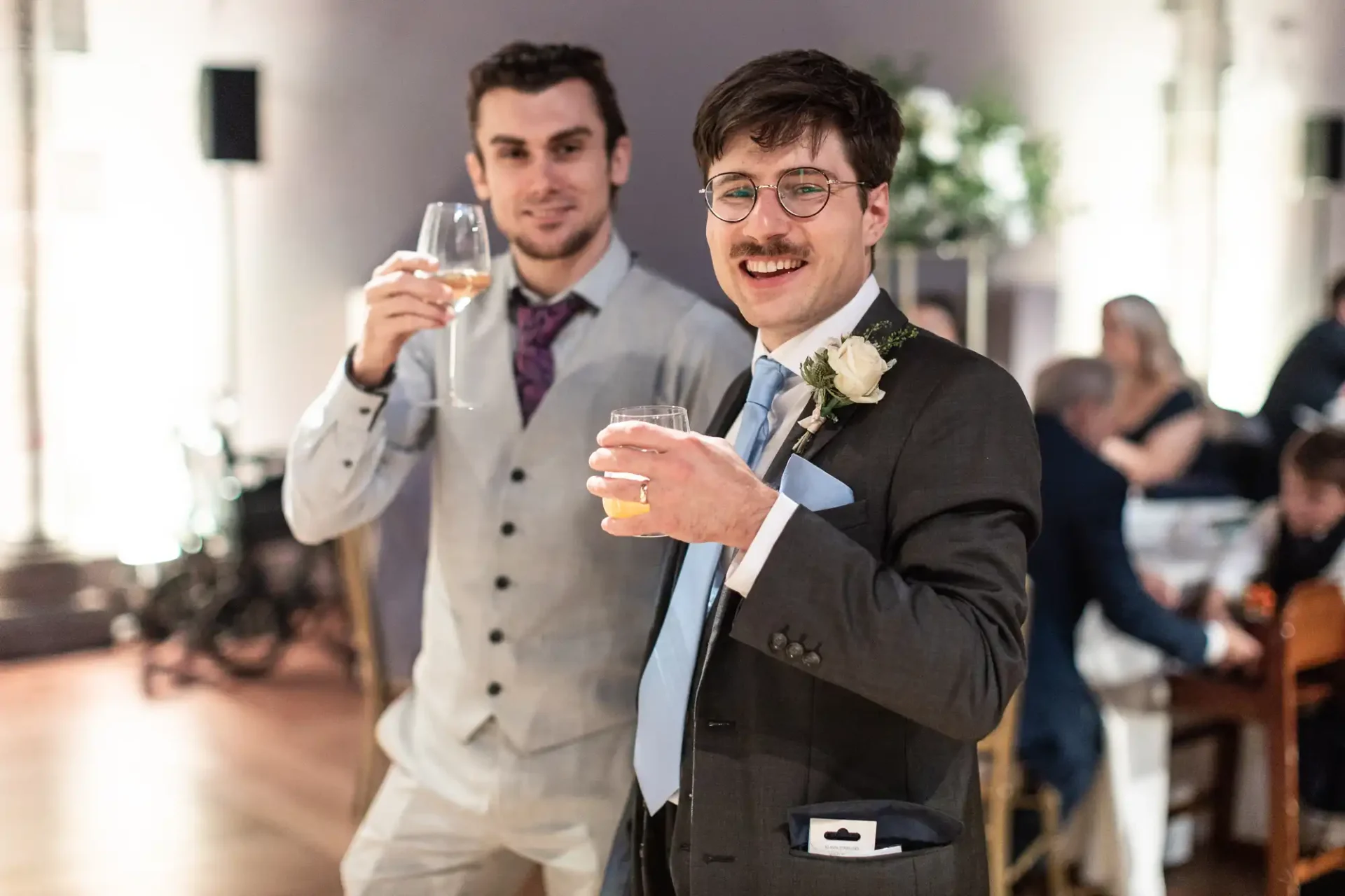 Two men in formal attire holding drinks and smiling at a gathering. One wears a gray suit, the other a dark suit with a boutonniere. Indoors with people and tables in the background.