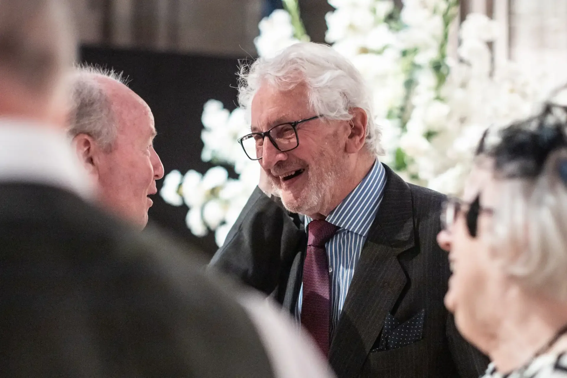 An elderly man in glasses and a suit smiles while engaging in conversation with others at a social gathering.