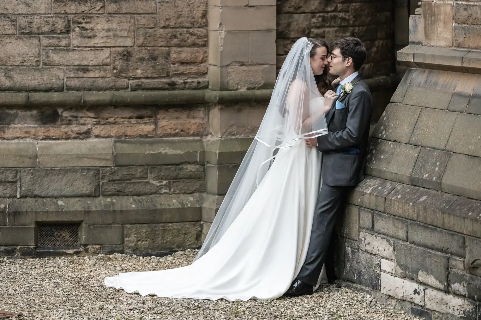 A bride in a white gown and veil stands with a groom in a gray suit, leaning against a stone building, sharing a moment.