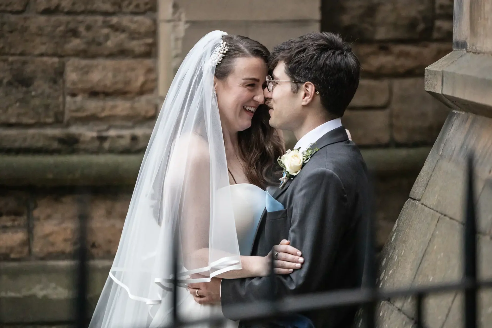 Bride and groom smiling at each other in front of a stone wall, with the bride wearing a veil and strapless dress, and the groom in a suit with a boutonniere.