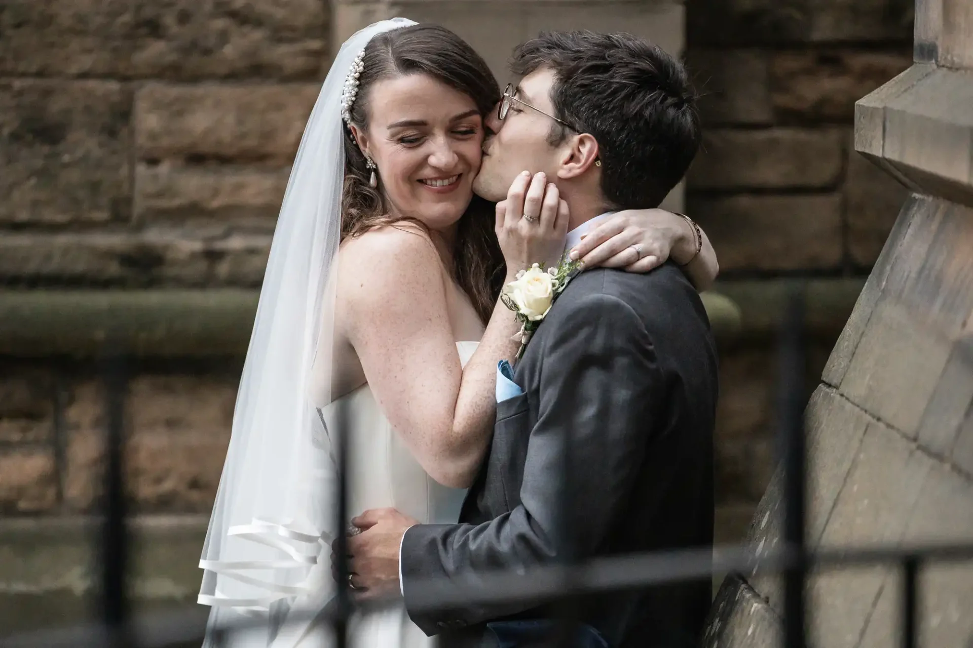 A bride and groom embrace near a stone wall. The groom kisses the bride's cheek, and both are smiling. The bride wears a white dress and veil, and the groom is in a dark suit with a boutonniere.