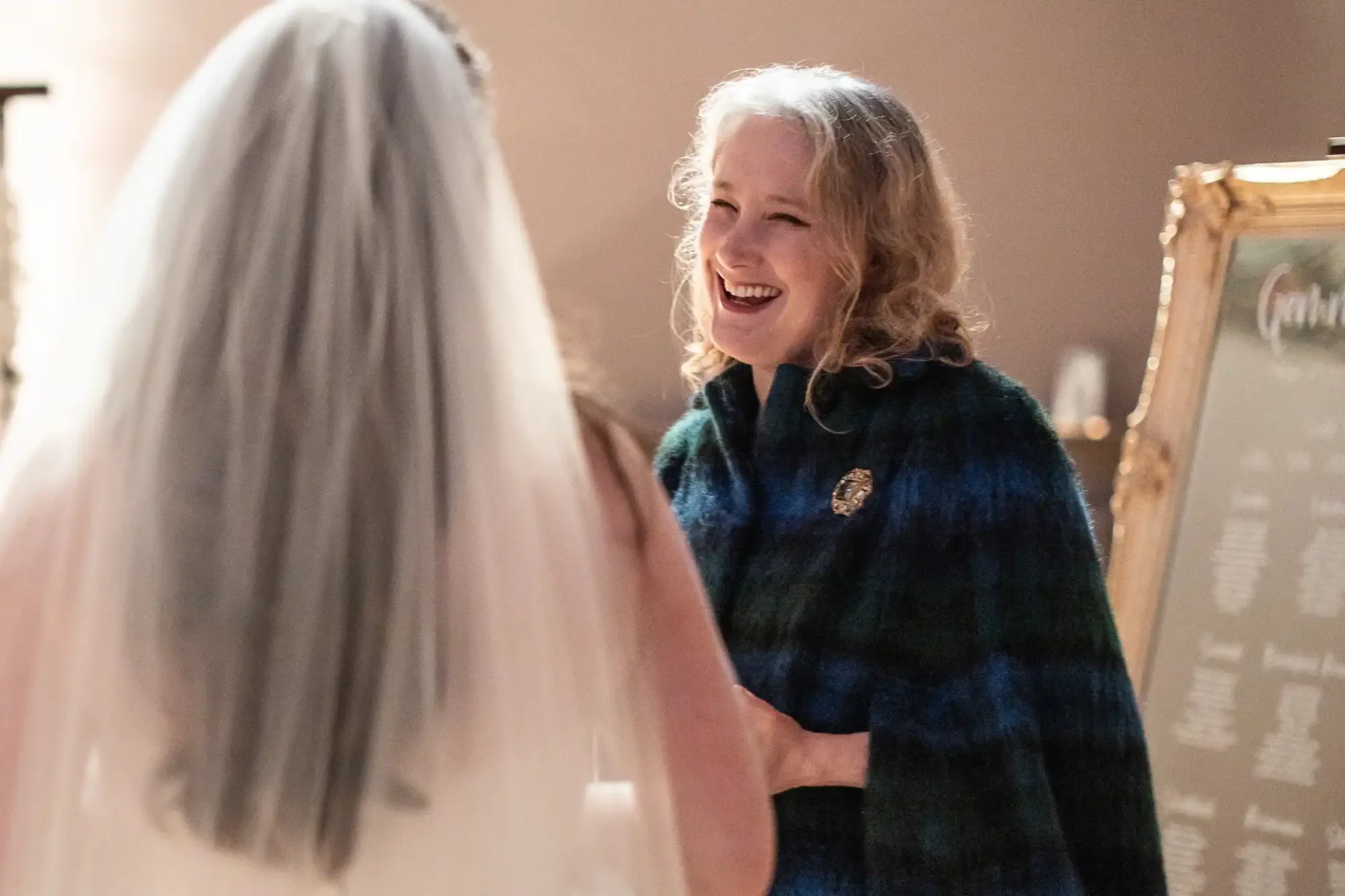 A woman in a wedding gown faces another woman with curly hair, wearing a dark shawl, and both are smiling indoors.