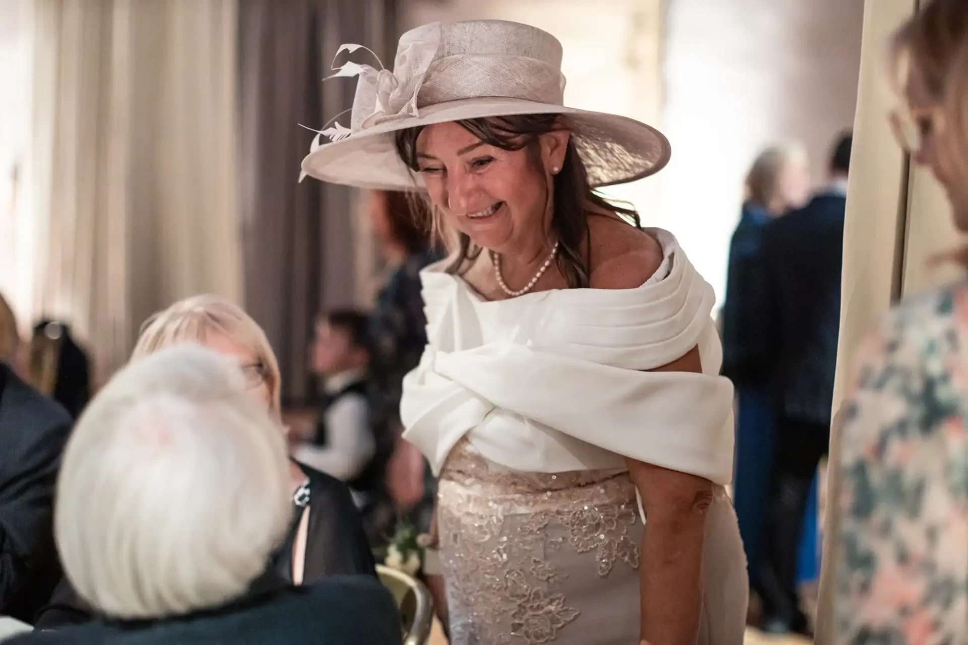 Woman in a decorative hat and elegant dress smiling while talking to seated guests at an indoor event.