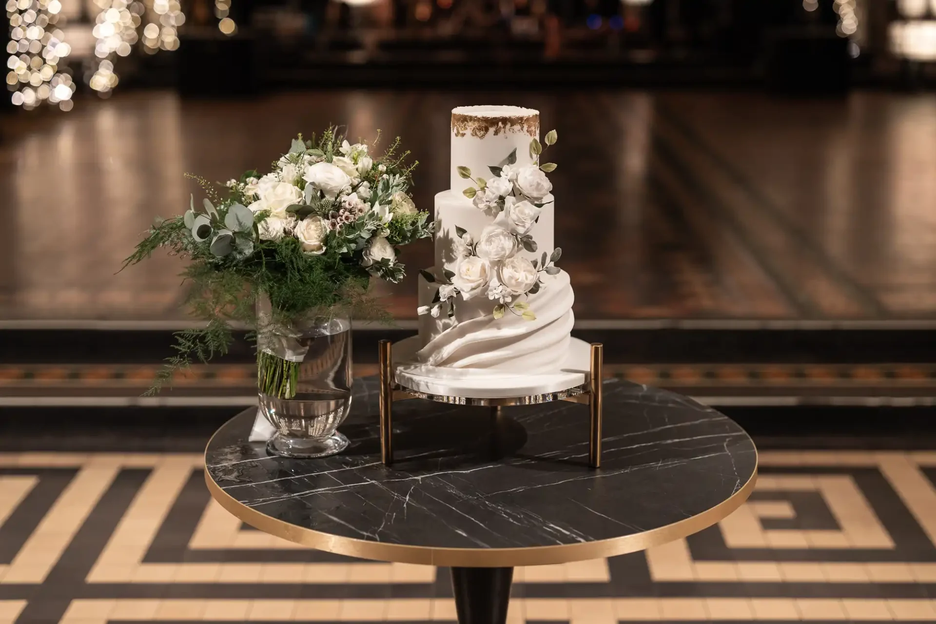 A white, tiered wedding cake with floral decorations sits on a black marble table next to a bouquet of white flowers in a glass vase.